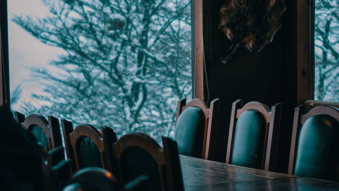 High-backed green leather chairs along a long wooden table in a lodge dining room, with snow-dusted conifers visible through a tall window