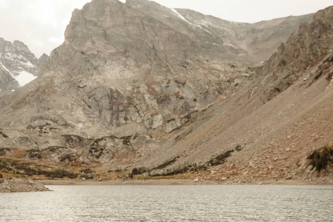 A bride in a long veil and a groom in a tan suit embrace on the rocky shore of a high alpine lake below bare mountain peaks.