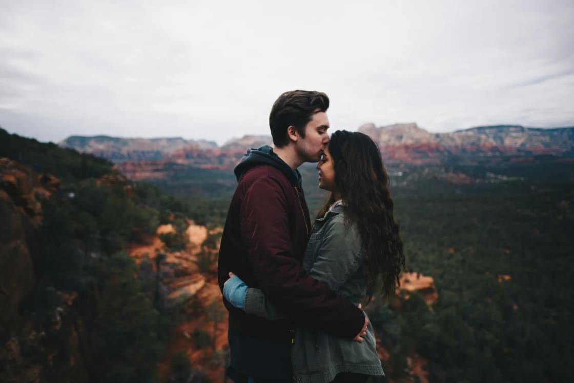A man kisses his fiancee's forehead during an engagement session on a cliff edge above red rock canyons and dark pines.