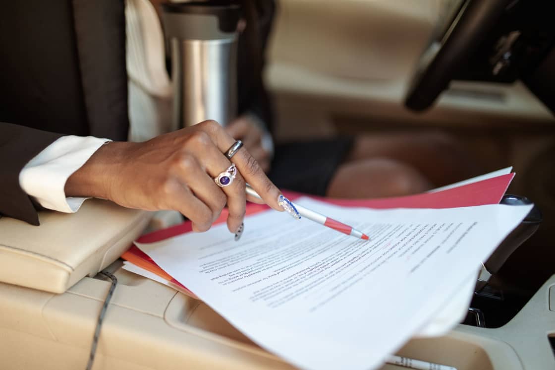 Una mano con bolígrafo revisa documentos impresos sobre la consola de un automóvil, con el volante al fondo