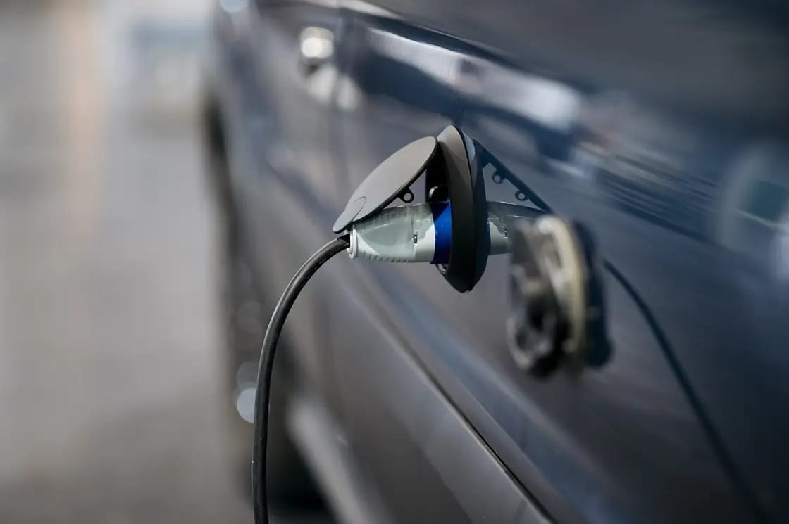A charging connector plugged into the open charge port of a dark gray electric car, the cable curving away out of frame