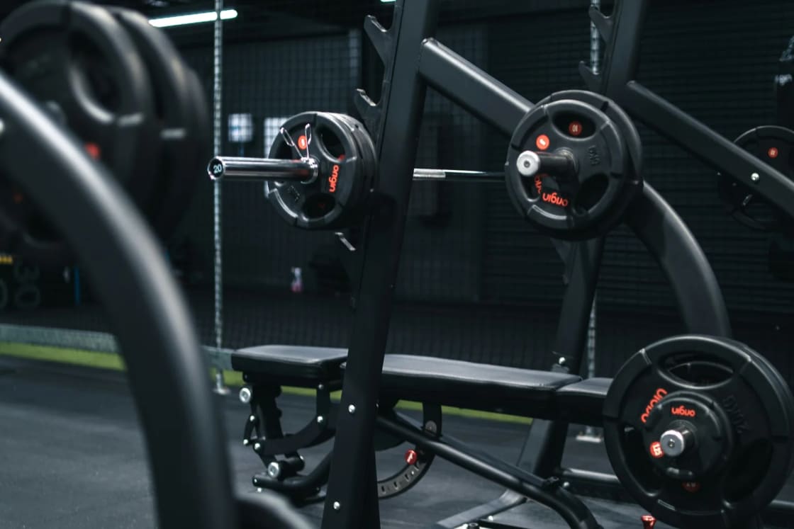 A dimly lit gym with a row of weight machines and loaded plates in the foreground and more equipment under strip lights behind