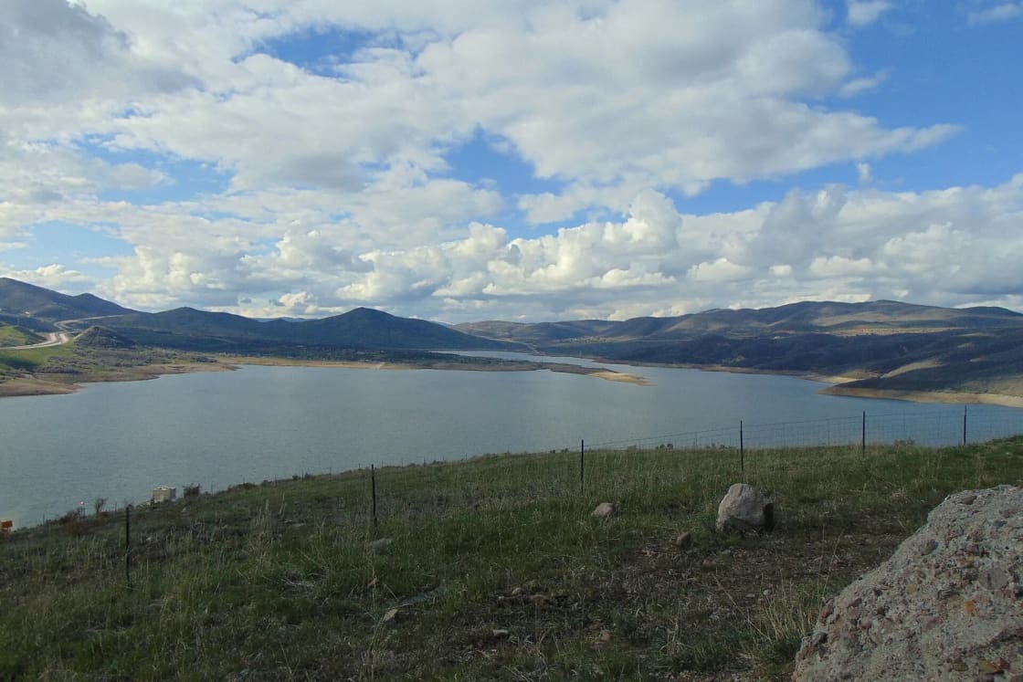 Jordanelle Reservoir seen from a grassy overlook, its blue water stretching between brown hills under a sky full of white clouds