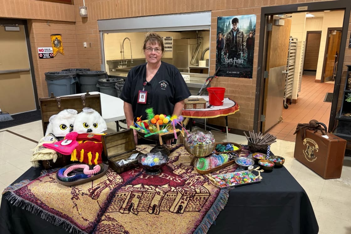 LaVon Vogl standing behind her Breakfast Games prize table, covered with a Marauder's Map blanket, Hedwig owl hats and bowls of prizes, in the Orchard Elementary cafeteria