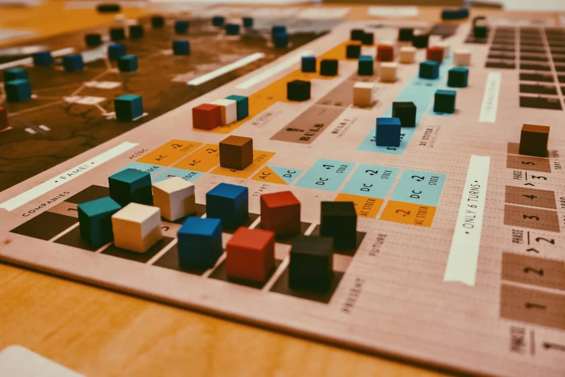 A board game in progress on a wooden table, colored wooden cubes scattered across the printed spaces of the game board.