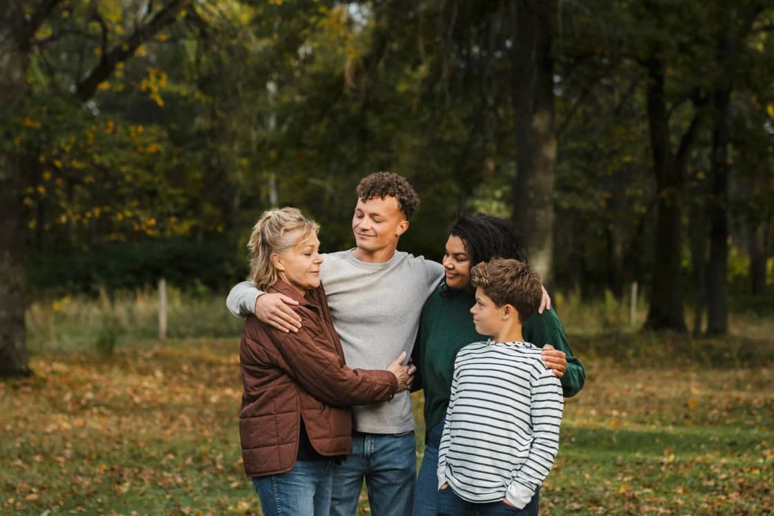 A family of four hugging and laughing together in a park in autumn, fallen leaves on the grass around them