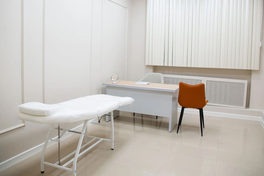 An empty consultation room with a padded examination table, a desk with an open notebook, and a single orange chair