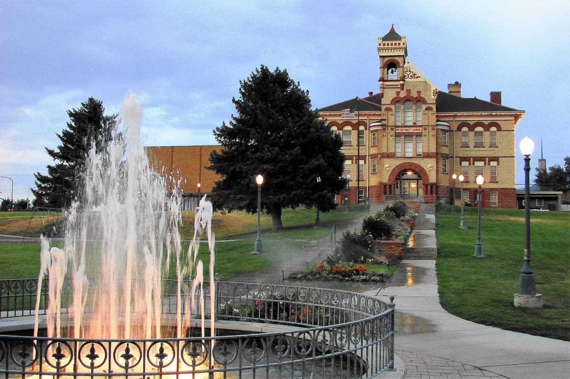 The Peteetneet Academy building in Payson, cream and red brick with a central bell tower, seen up a lawn behind a splashing fountain ringed by an iron railing, with lampposts and flower beds under a blue sky