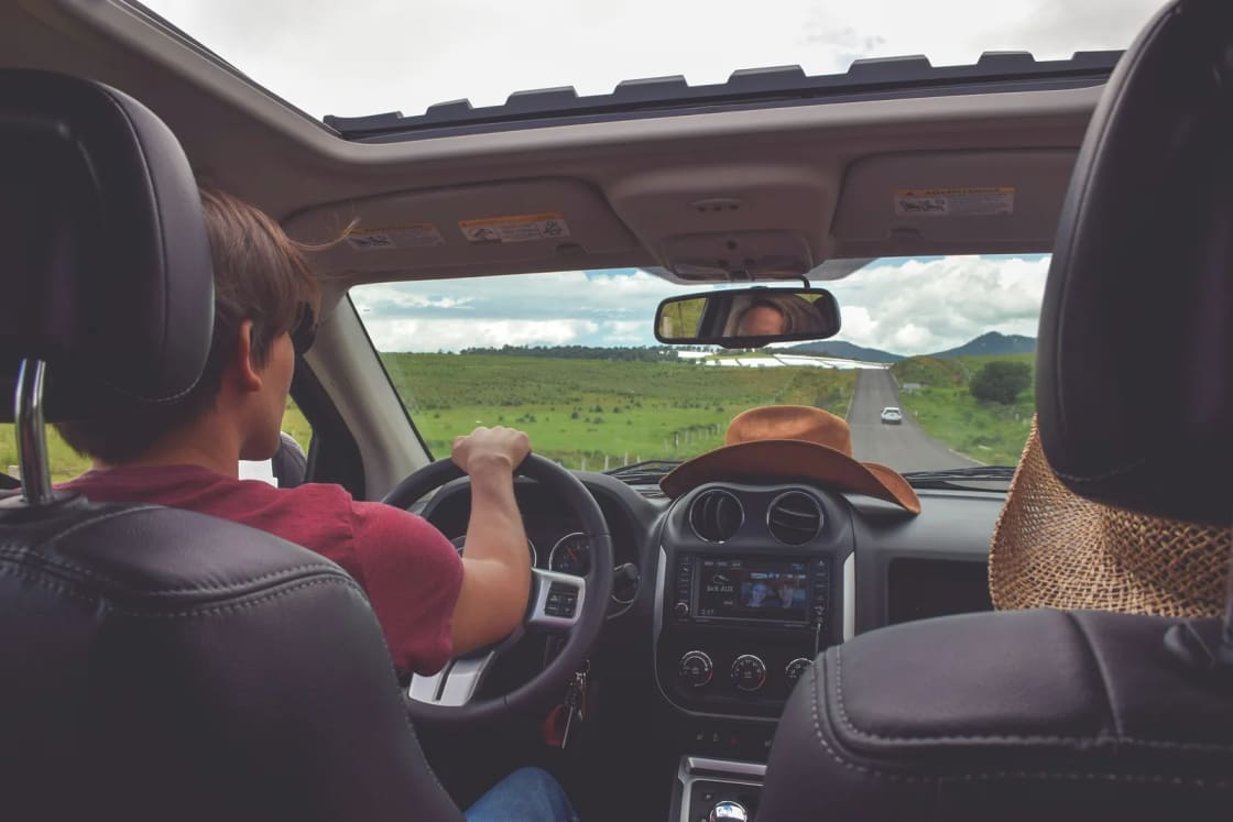View from the back seat of a car on a road trip, the driver heading down a two-lane road through green hills