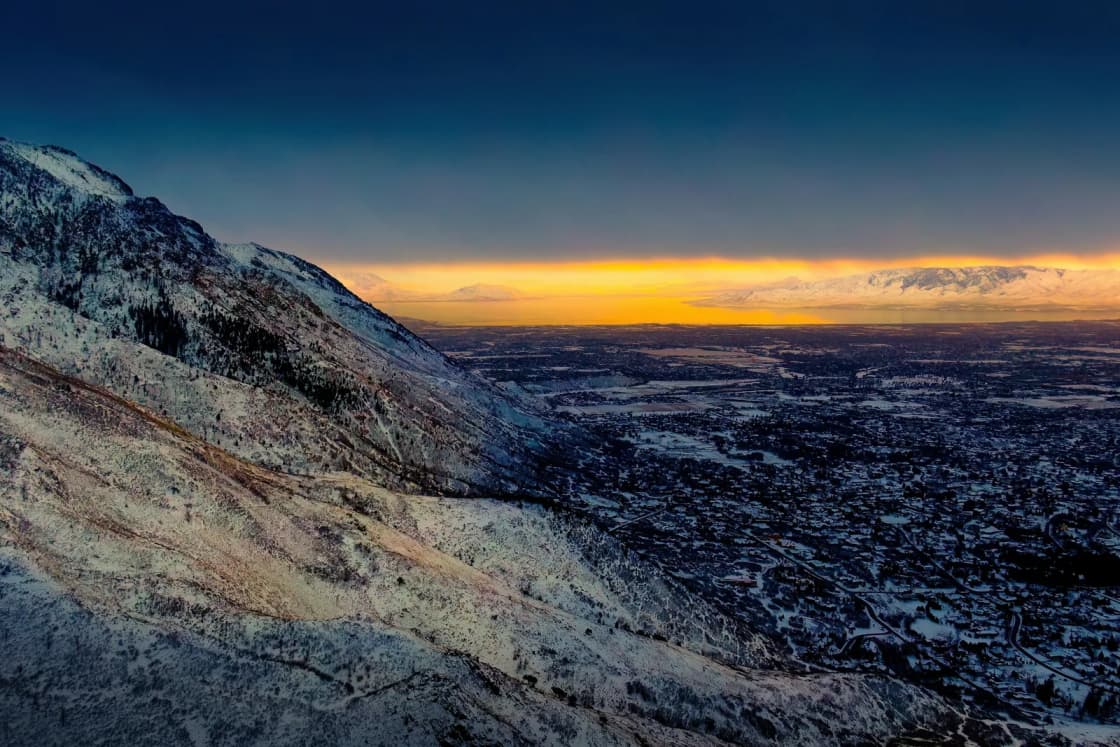 A deep orange winter sunset glows along the horizon beyond a dark valley, seen from a rocky, wind-scoured mountain slope.