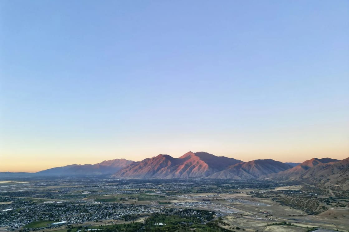 Early light warms a mountain range above a wide, green valley floor of neighborhoods and fields on a clear summer morning.