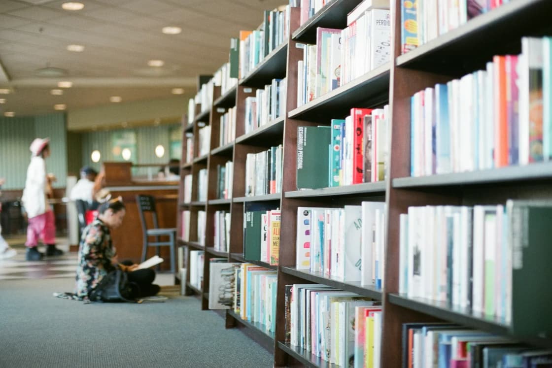 Tall shelves of books in a quiet public library, a reader sitting on the carpet in the aisle with an open book