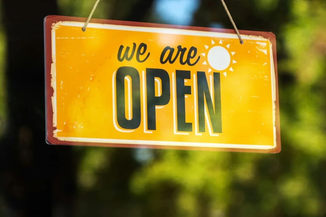 A yellow we are open sign hanging by a string in a shop window, green trees blurred in the background