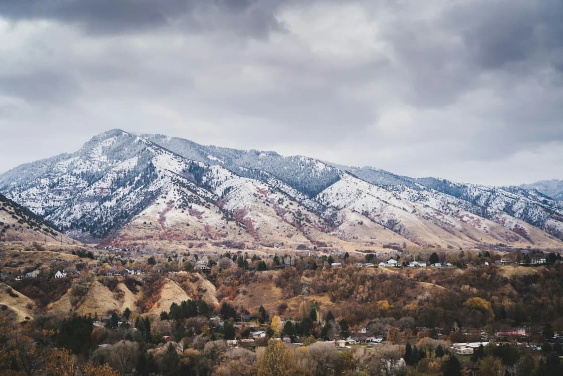 The first snow of the season dusts a mountain range above a town and autumn-brown foothills under heavy gray clouds.