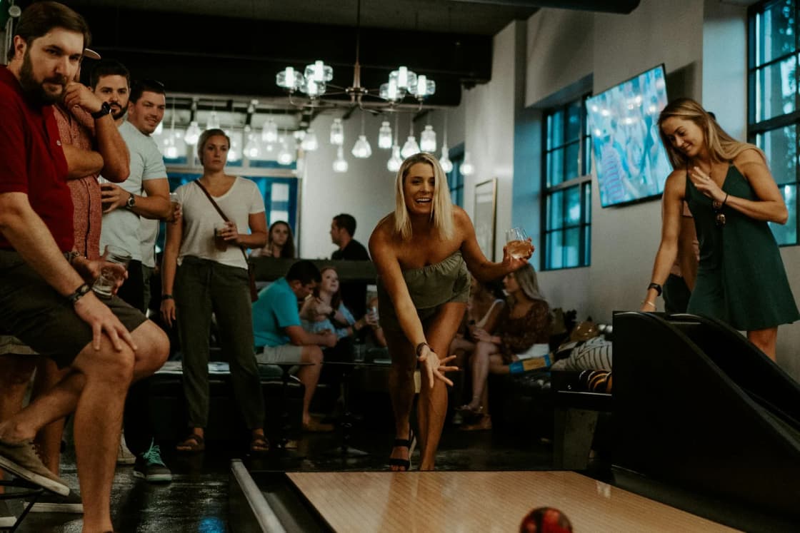 A bowler releases a ball down a polished lane while a group of friends watches from behind in a dim, string-lit bowling alley.
