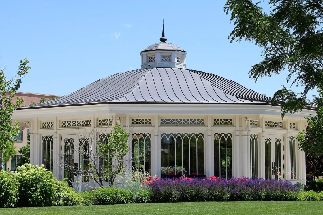 A white octagonal garden pavilion with tall windows and a metal roof among flower beds and lawn on the Provo City Center Temple grounds.