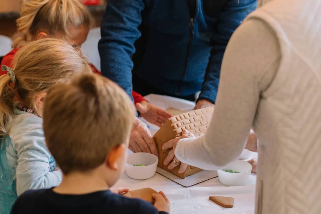 Children gathered at a kitchen table with two adults, building and decorating a gingerbread house together