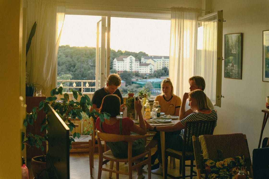 A group of young adults sharing dinner around a table in a sunny apartment, a balcony door open to green hills