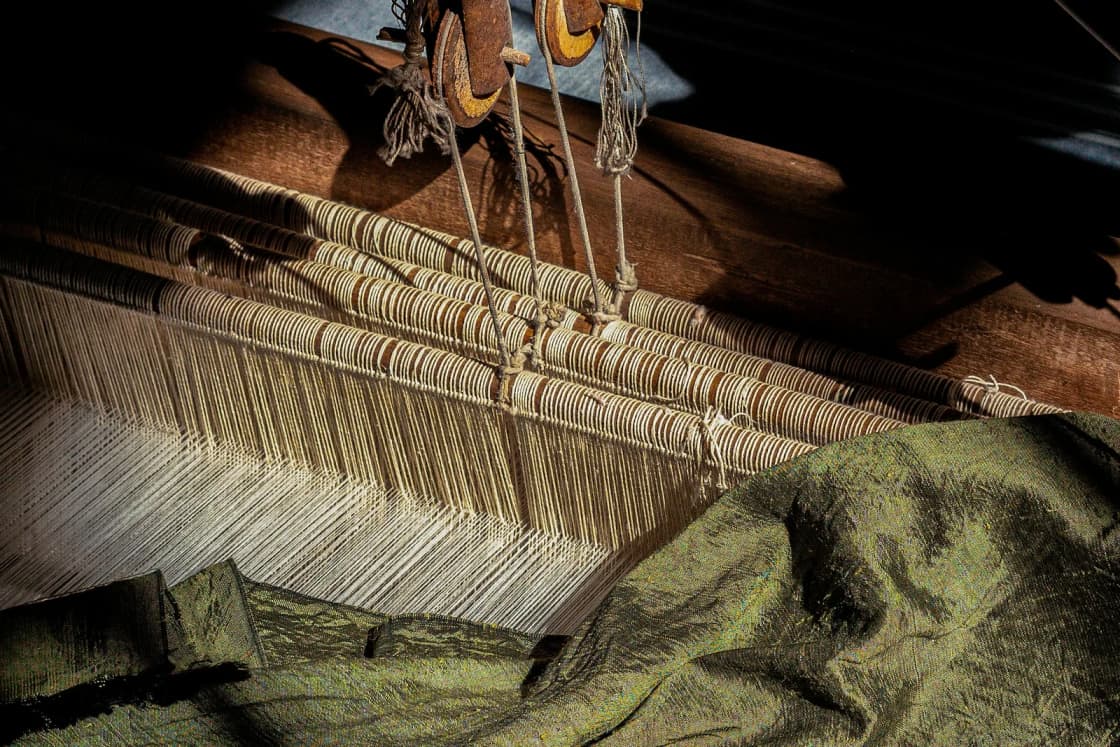 Close-up of a wooden floor loom mid-weave, with taut warp threads and a length of green cloth gathered on the beam.