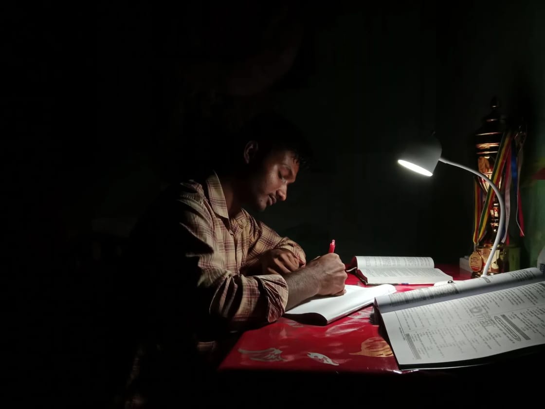 A student writing in a notebook at a desk lit only by an angled desk lamp, with an open textbook and a workbook beside him in a dark room