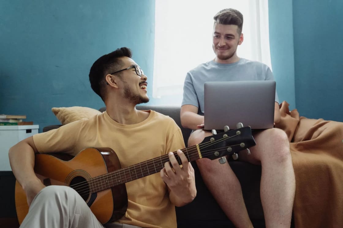 Two young men on a sofa in a shared living room, one playing an acoustic guitar and the other working on a laptop