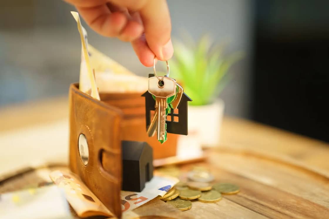 Close-up of hand holding a house key with a wallet and coins, symbolizing real estate investment