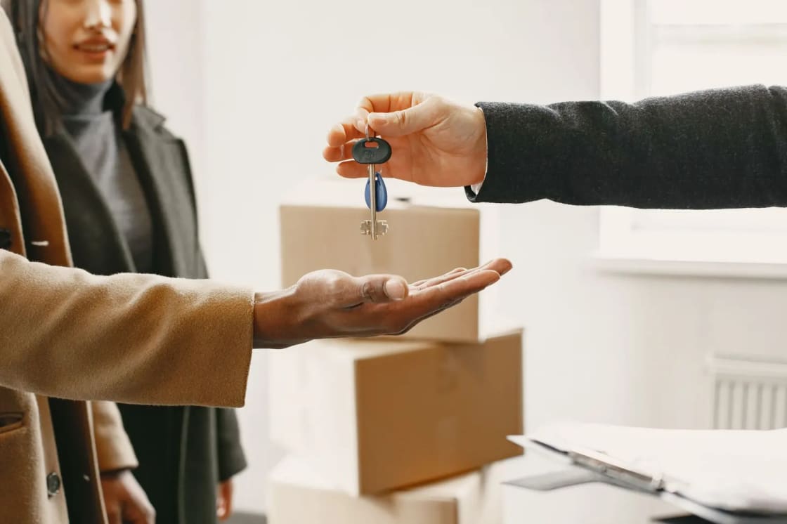 One person handing a set of keys to another's open palm in a room stacked with cardboard moving boxes