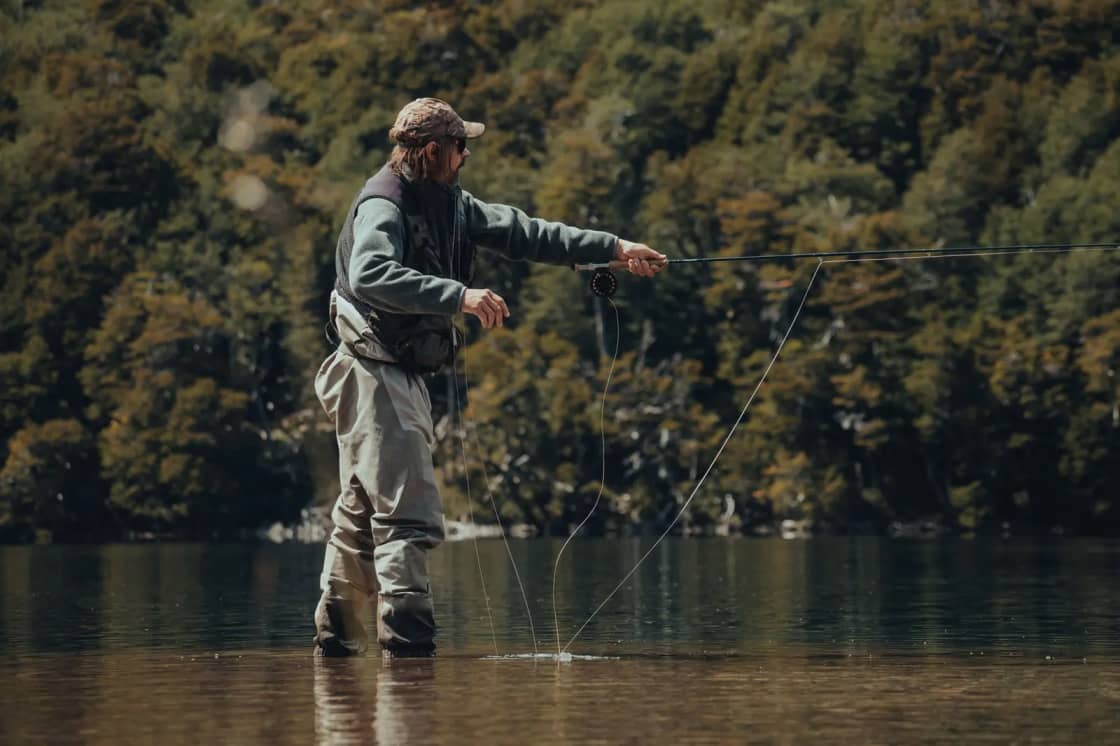 An angler in chest waders casting a fly rod while standing in shallow, still water below a wooded shoreline
