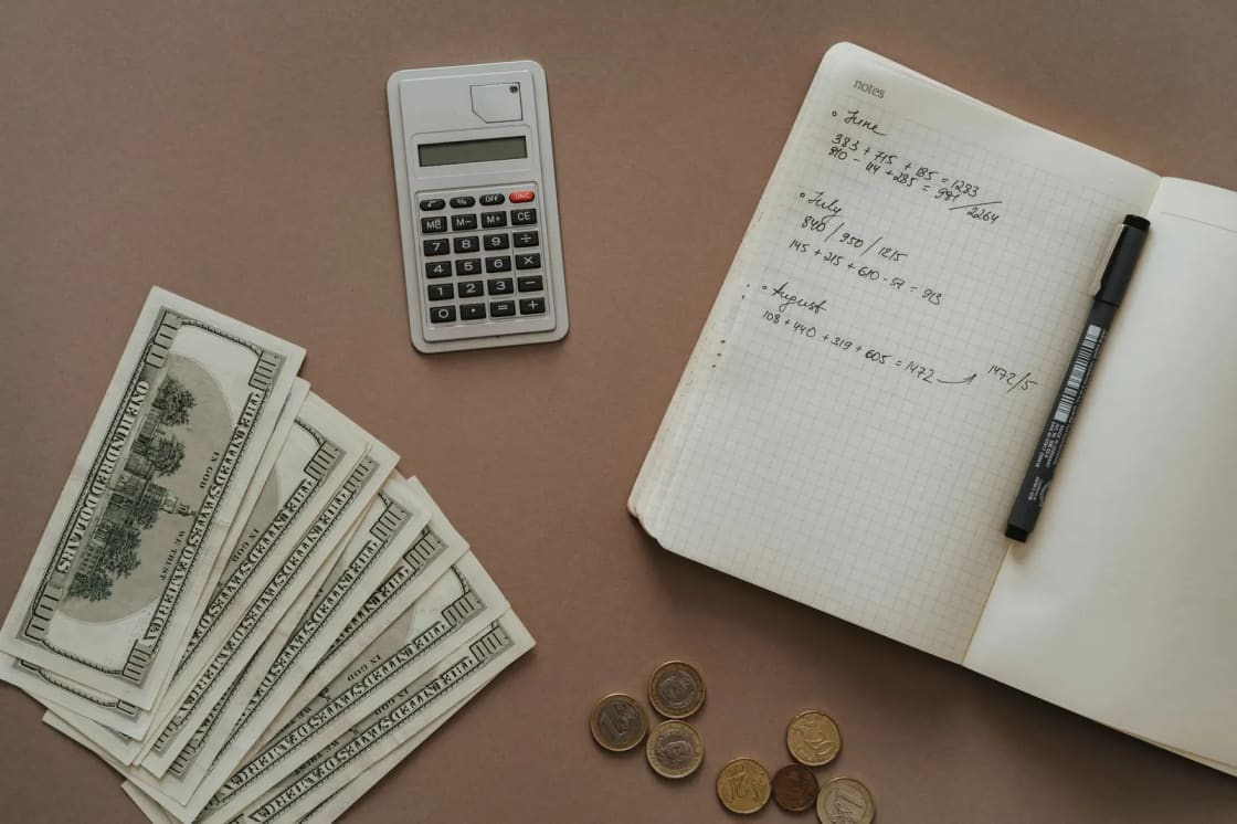 US banknotes, loose coins and a calculator laid beside an open grid-paper notebook filled with handwritten monthly calculations
