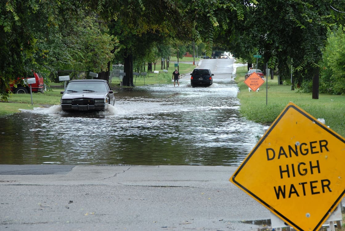 A car driving through floodwater covering a residential street, past a yellow DANGER HIGH WATER sign and a stalled vehicle