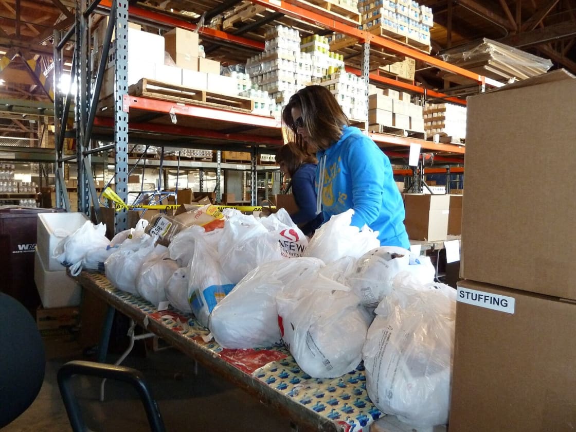 Volunteers packing bagged groceries at long tables in a food bank warehouse