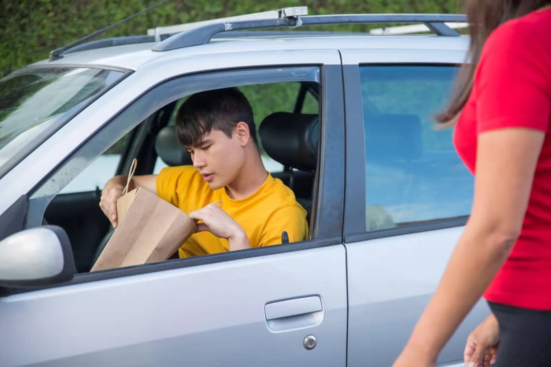 A driver handing a paper takeout bag out of a car window to a waiting customer