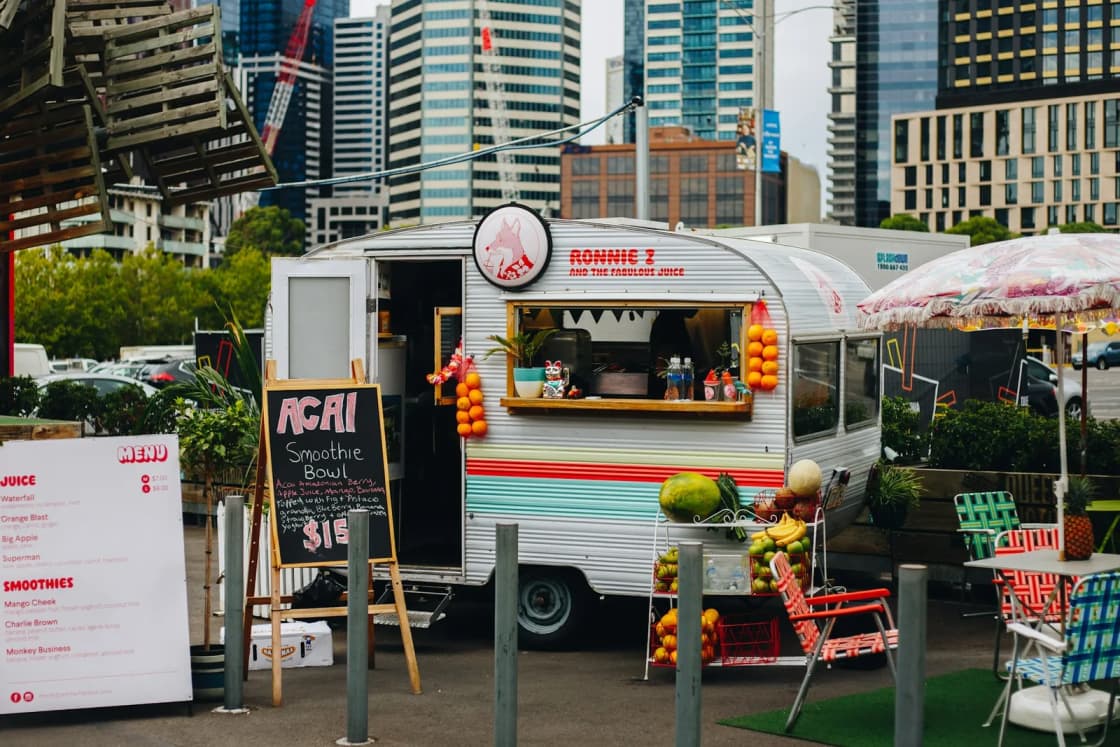 A small white vintage food trailer with a striped awning, chalkboard menus and patio chairs set out on artificial turf under city towers