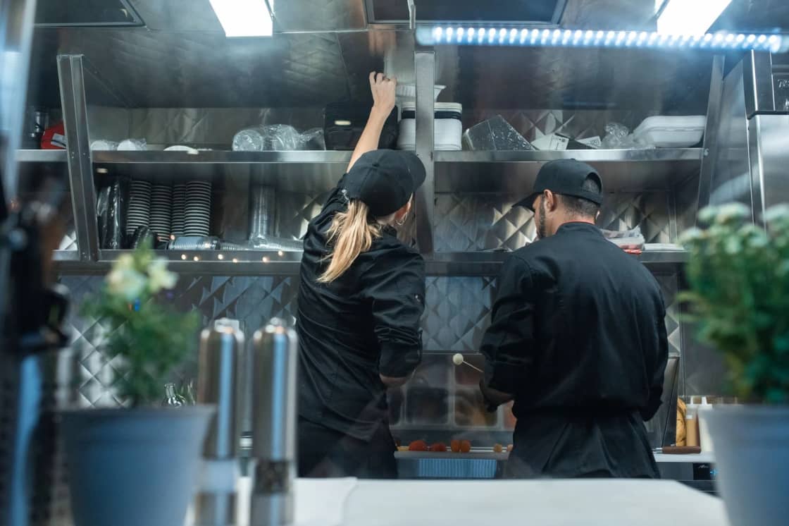 Two cooks in black jackets and caps working at a stainless steel prep counter in a commercial kitchen