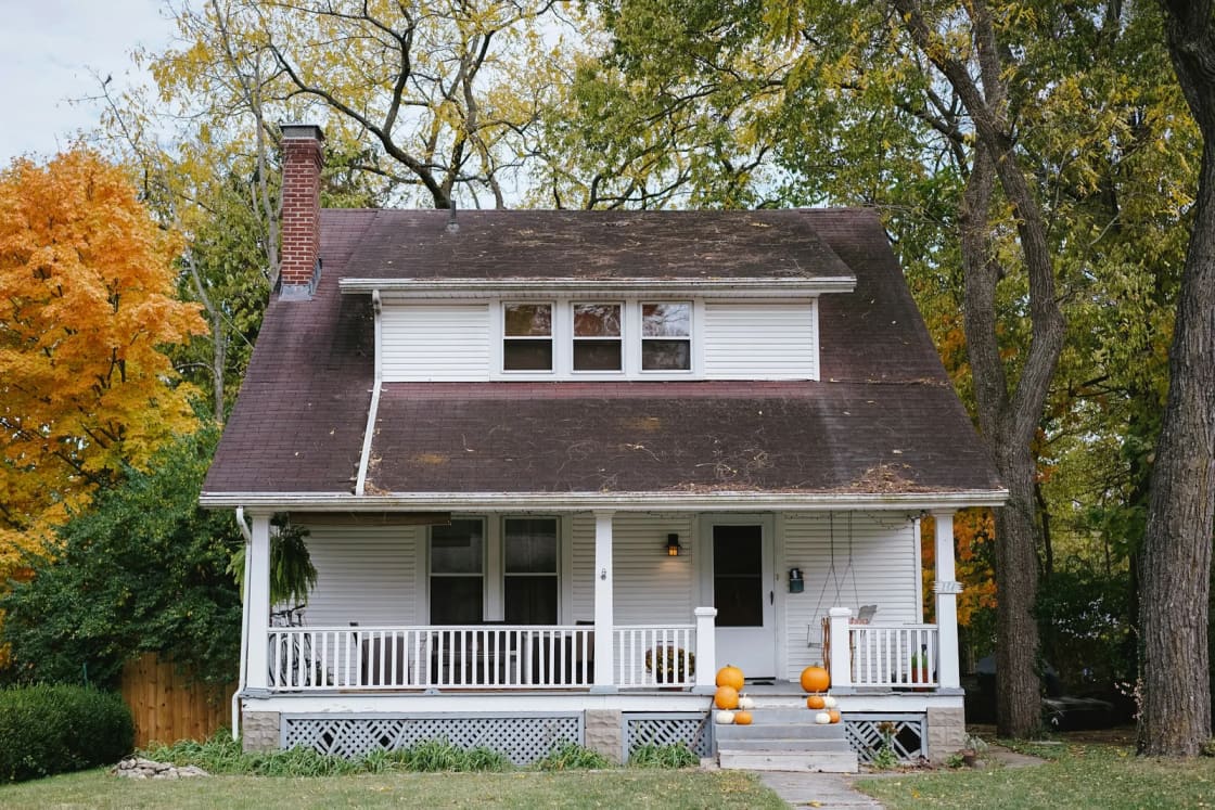 A white bungalow with a dark roof, brick chimney and front porch decorated with pumpkins, framed by autumn trees with orange leaves