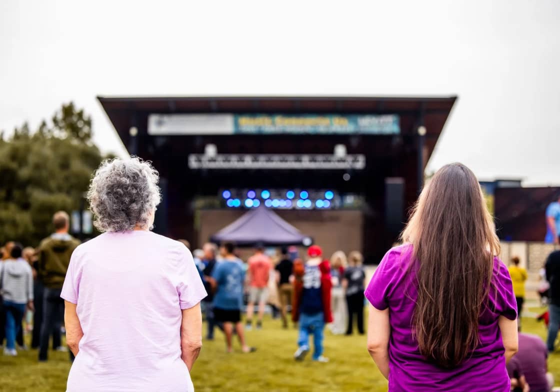 Two women seen from behind on the grass at an outdoor concert, looking toward a covered stage with blue lights and a crowd gathered in front of it