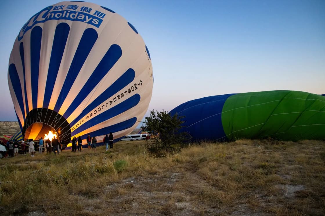 A hot air balloon standing upright at dawn with its burner lit, a second envelope still lying inflated on the grass beside it