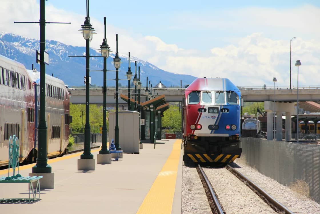 A red, white and blue FrontRunner locomotive waits at the Ogden station platform beside its passenger cars, with snowy peaks behind.