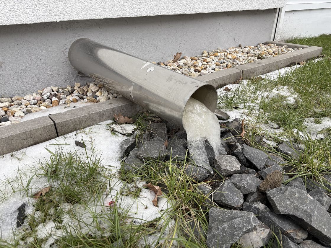 A plug of ice frozen in the mouth of a drainpipe at the base of a house, with thin snow on the ground