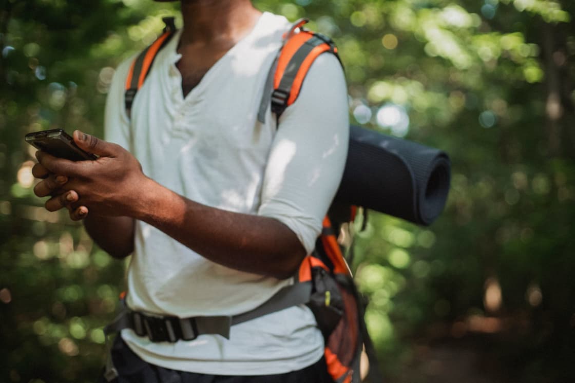 A hiker in a pale shirt with an orange backpack and rolled sleeping mat standing on a sunlit forest trail, looking down at a phone held in one hand