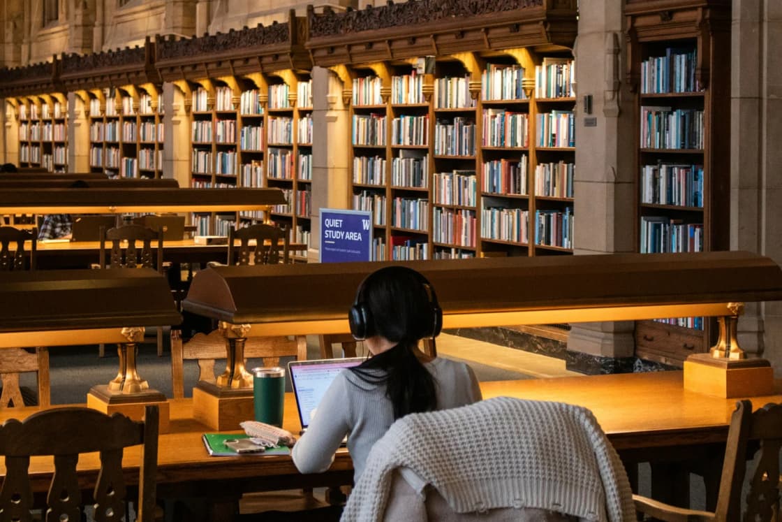 A student in headphones works on a laptop at a long reading-room table in a grand university library lined with bookshelves.