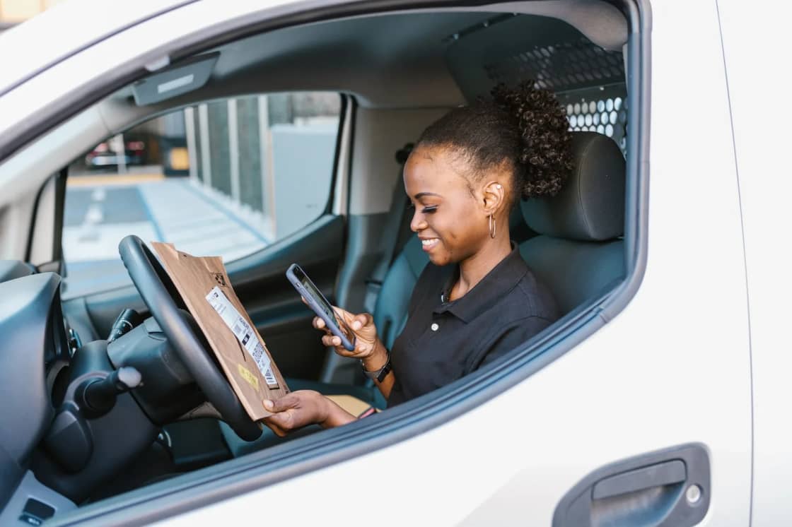 A delivery driver at the wheel of a white van, looking down at a phone held above a clipboard on her lap