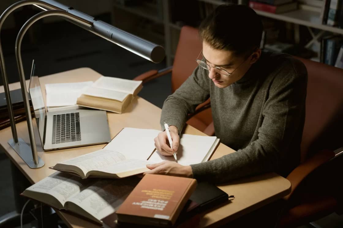 A student in glasses writing longhand in a bound notebook under an angled desk lamp, with a laptop and several open hardback books spread across the desk