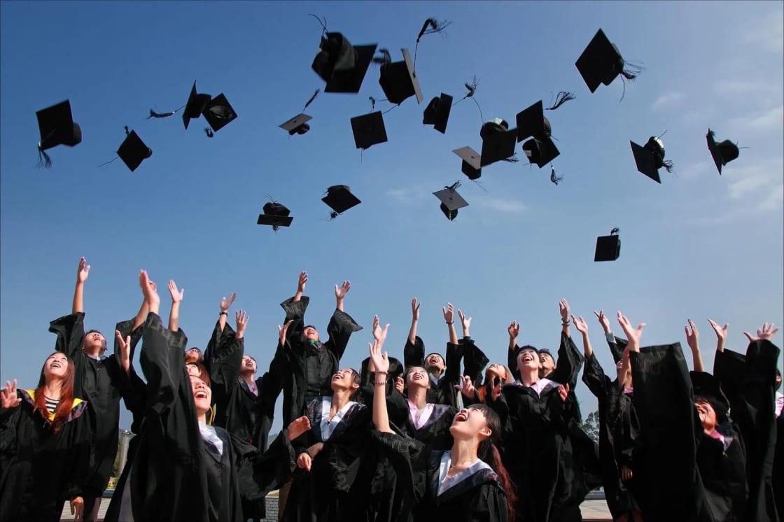 Graduates in black gowns with their arms raised, throwing mortarboard caps into a blue sky