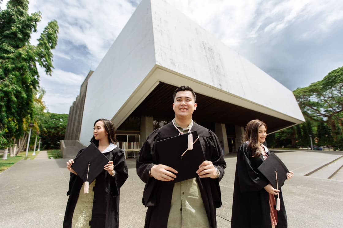 Three graduates in caps and gowns holding diplomas outside a modern concrete building