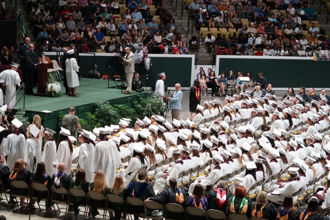 Rows of graduates in white caps and gowns seated in an arena, with a draped stage and tiered spectator seating behind them