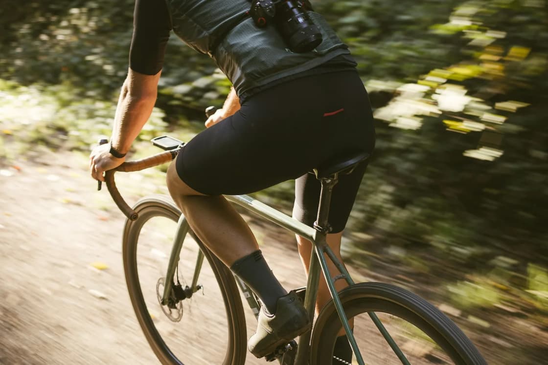A cyclist seen from behind riding a drop-bar bike fast down a dirt road through sunlit forest, the background blurred with speed