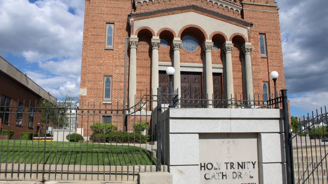 The arched brick portico of Holy Trinity Greek Orthodox Cathedral in Salt Lake City, with columns, lamps and an iron gate, and the words Holy Trinity Cathedral carved into the stone wall beside the steps