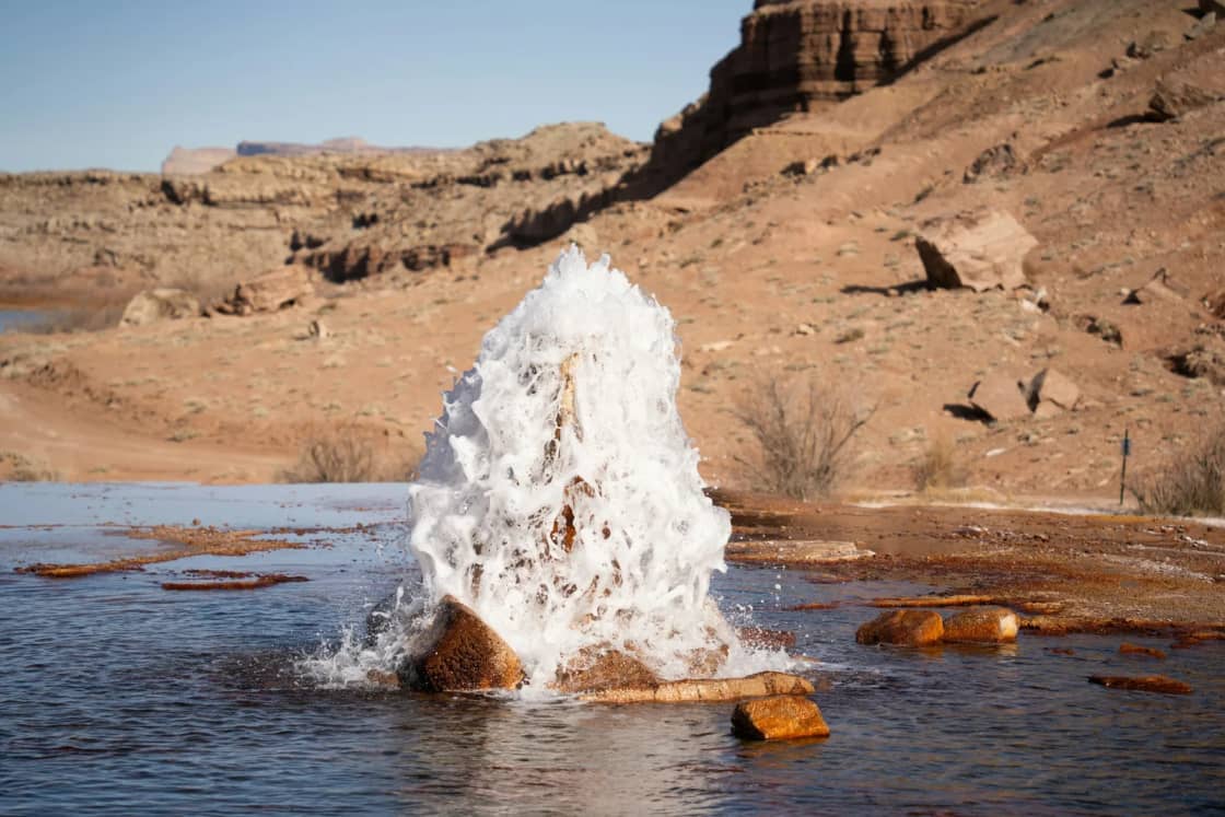 Crystal Geyser erupting a white column of water beside rust-orange mineral deposits near Green River, with sandstone ledges behind.