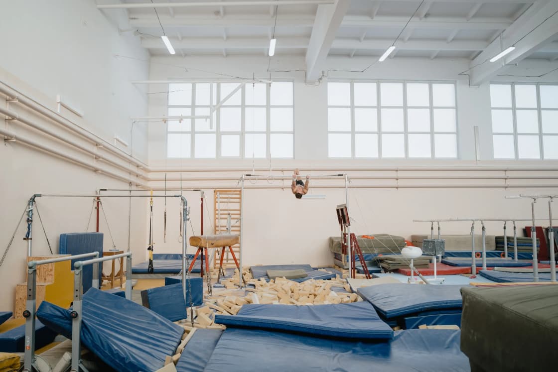 A bright gymnastics gym with blue mats, a foam pit and wall bars under high windows